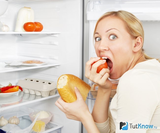 Девушка ест помидор и хлеб возле холодильника