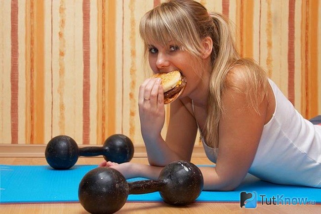Девушка ест гамбургер возле гантелей