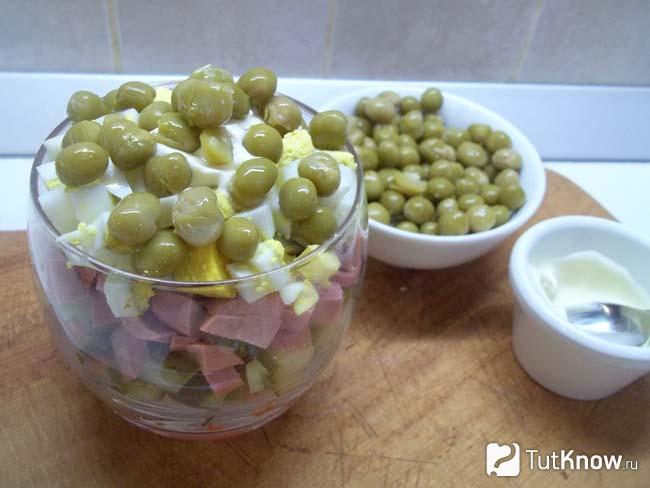 Готовый салат в стакане с сосисками, морковью, огурцом и яйцами присыпан зеленым горошком